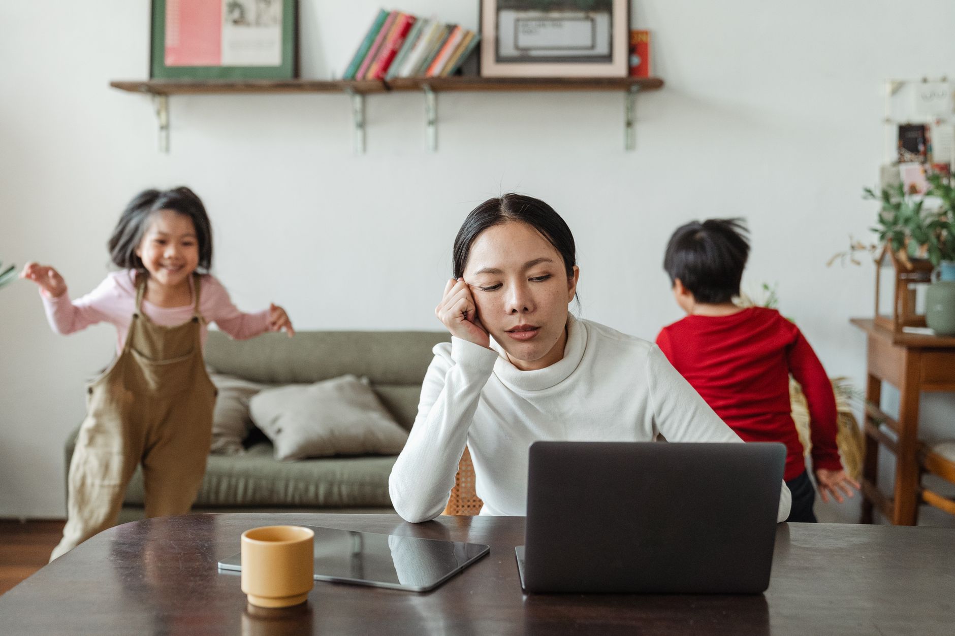 Zmęczona mama pracuje przy laptopie, dzieci hałaśliwie bawią się obok